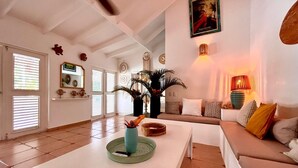 A spacious living area with a white sofa, a coffee table, and a decorative vase. There are two windows with blinds, a wall-mounted lamp, and a framed picture.