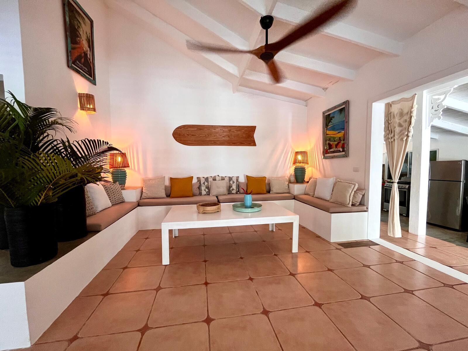 Una sala de estar con sofá blanco, una mesa de centro, ventilador de techo y cocina visible a través de un vano abierto.