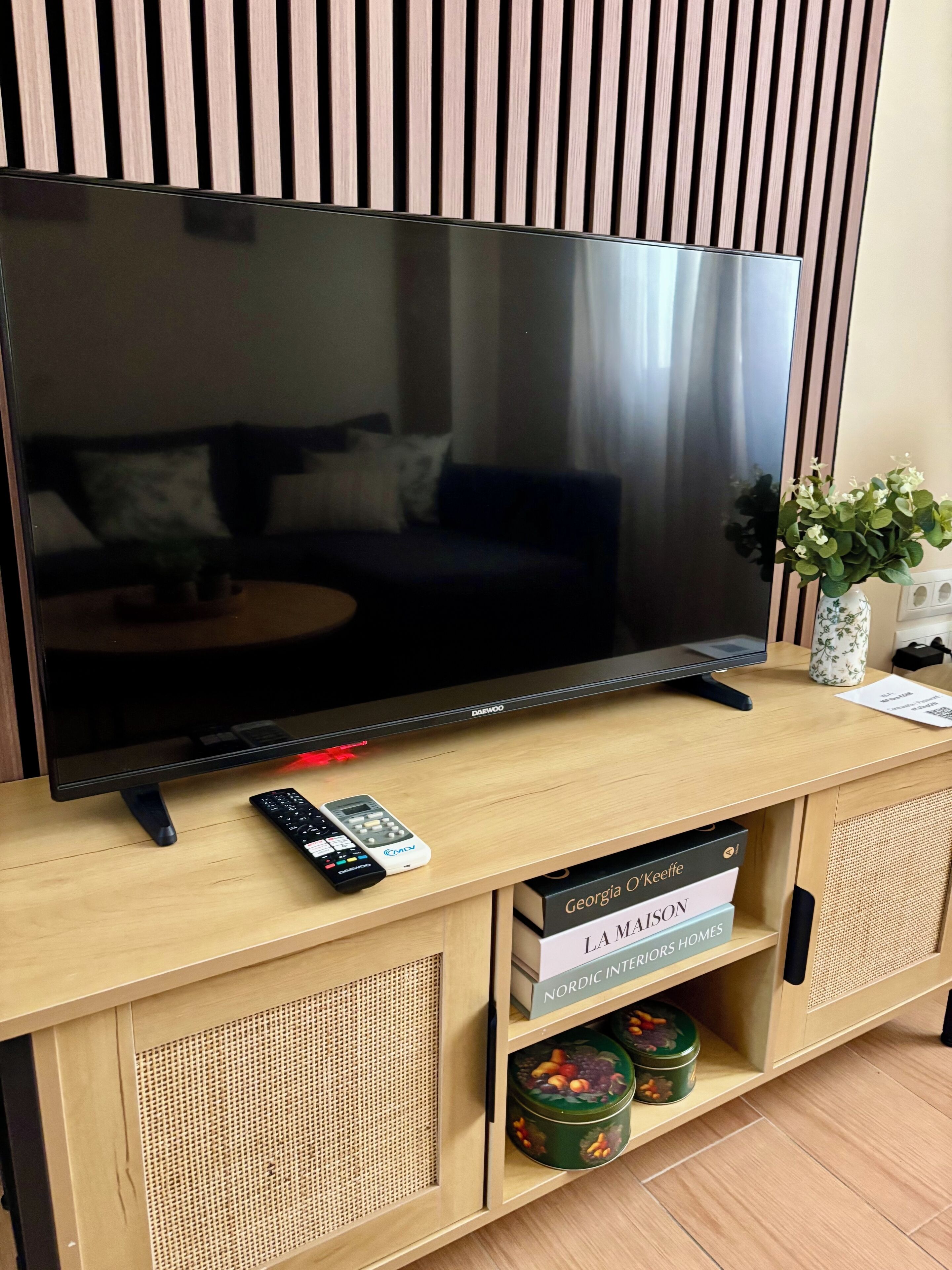 Un téléviseur à écran plat posé sur un meuble télé en bois, avec des télécommandes à proximité, un vase rempli de fleurs et des boîtes de rangement étiquetées « Georgia O’Keeffe » et « Intérieurs nordiques ».