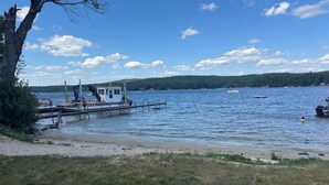 Un muelle junto al lago con un bote pequeño y una cabaña, rodeado de zonas verdes y una colina boscosa.