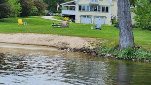 Una casa grande con un muelle que se adentra en un lago tranquilo, rodeada de vegetación exuberante y una iglesia al fondo.