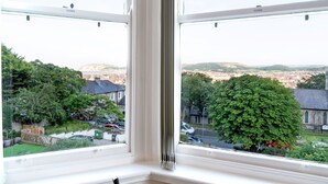 Une vue depuis une fenêtre dont les stores sont fermés, donnant sur un quartier résidentiel avec des maisons, des arbres et une colline au loin.