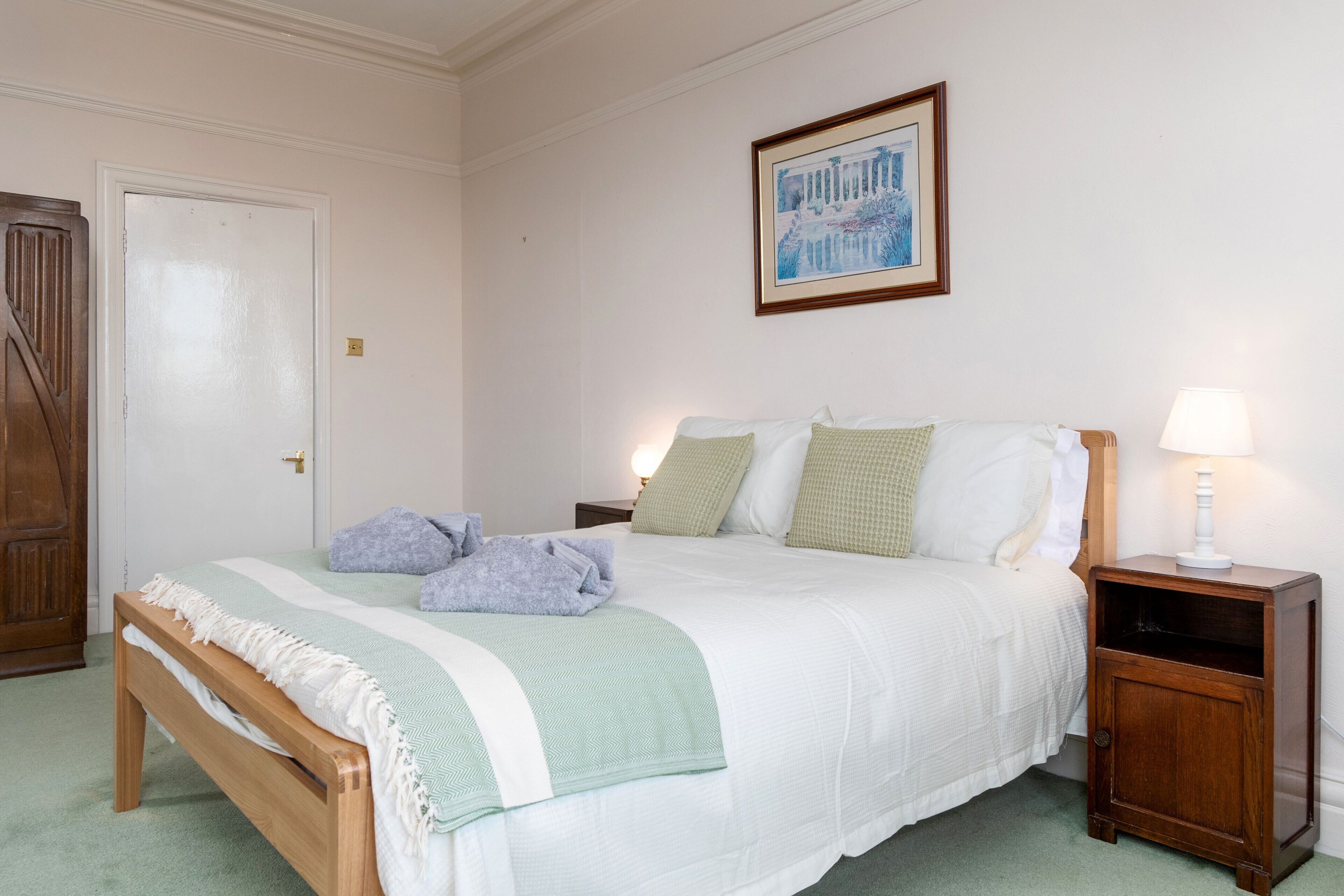 Una cama bien hecha con cabecera de madera, una mesita de noche con lámpara y un cuadro enmarcado en la pared.