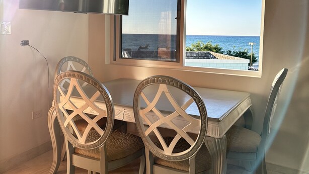 A dining area with a table and chairs, a window with a view of the sea, and a ceiling light. - Beach Condo in Negril Jamaica (Negril)