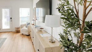 Ein modernes Wohnzimmer mit Holzboden, einem großen Flachbildfernseher, einem weißen Sofa, einem Beistelltisch mit Lampe und einer Zimmerpflanze.