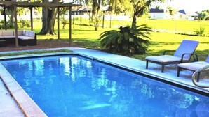 Une piscine rectangulaire bordée de carreaux, entourée d’une pelouse agrémentée de quelques arbres et d’un espace de détente couvert, équipé de coussins.