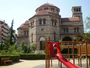 A stone building with a red-tiled roof and arched windows, surrounded by greenery and a playground with a red slide.