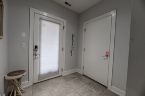 A hallway with two white doors, one with a closed window and the other with a closed door. There is a small wooden stool and a wall-mounted branch decoration. - Marcy Condo (Omaha)