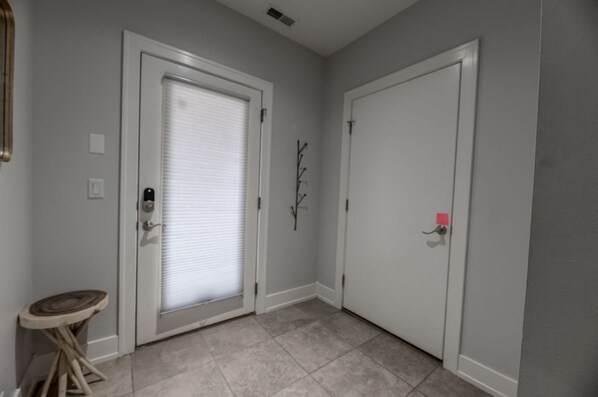 A hallway with two white doors, one with a closed window and the other with a closed door. There is a small wooden stool and a wall-mounted branch decoration.
