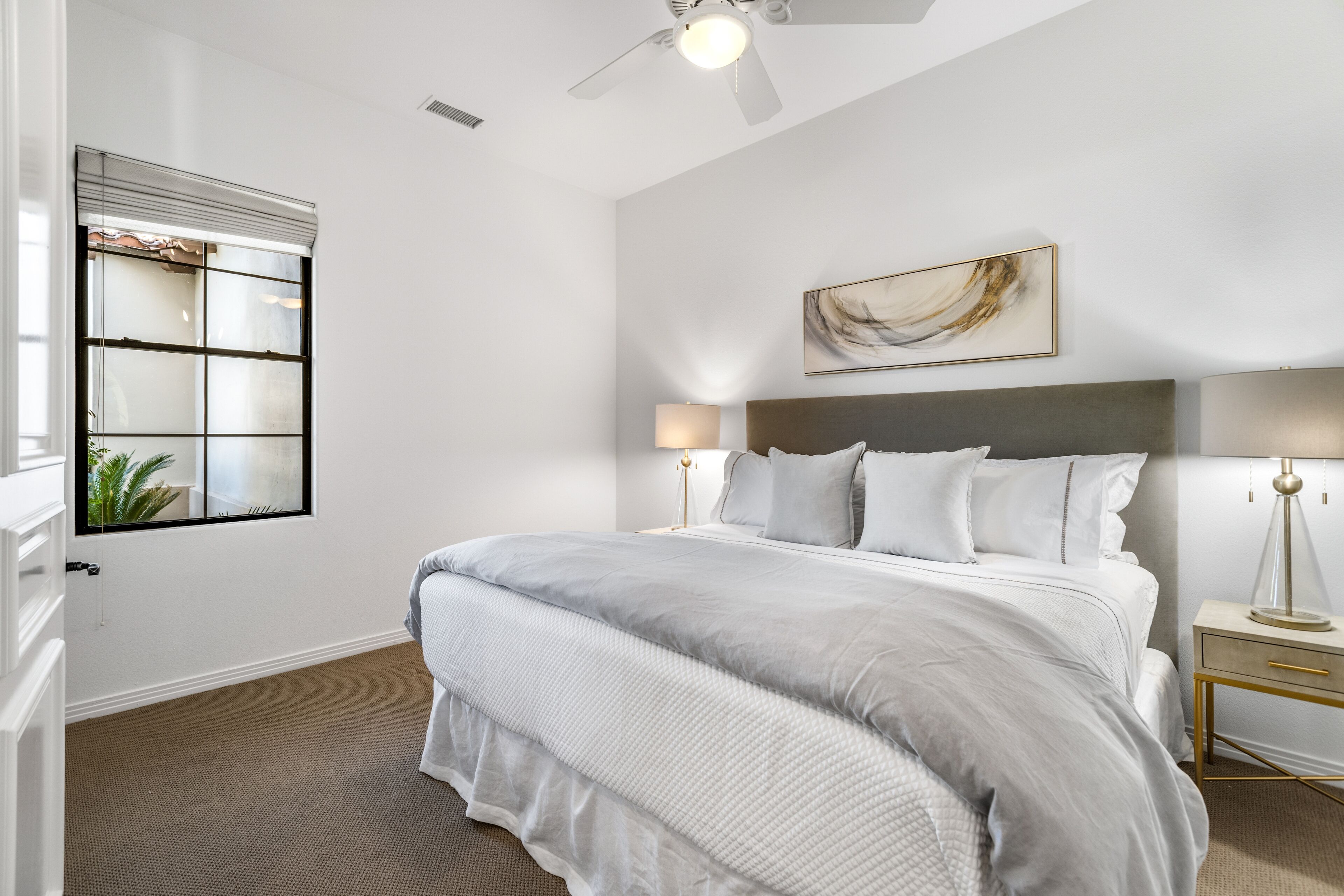 A bedroom with a large bed, a ceiling fan, a window with blinds, and a bedside table with a lamp.