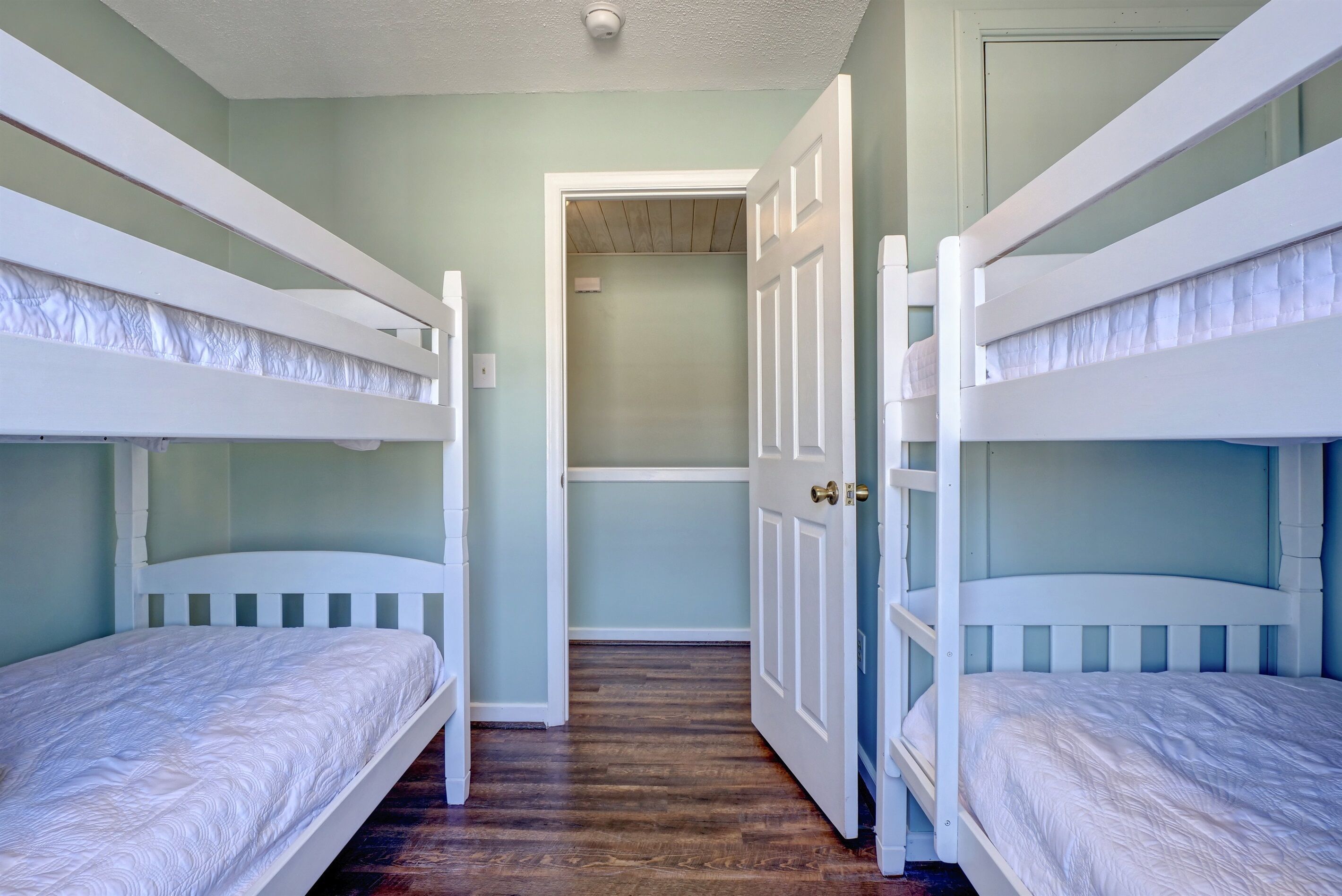 A room with bunk beds, a wooden floor, and a door leading to another room.