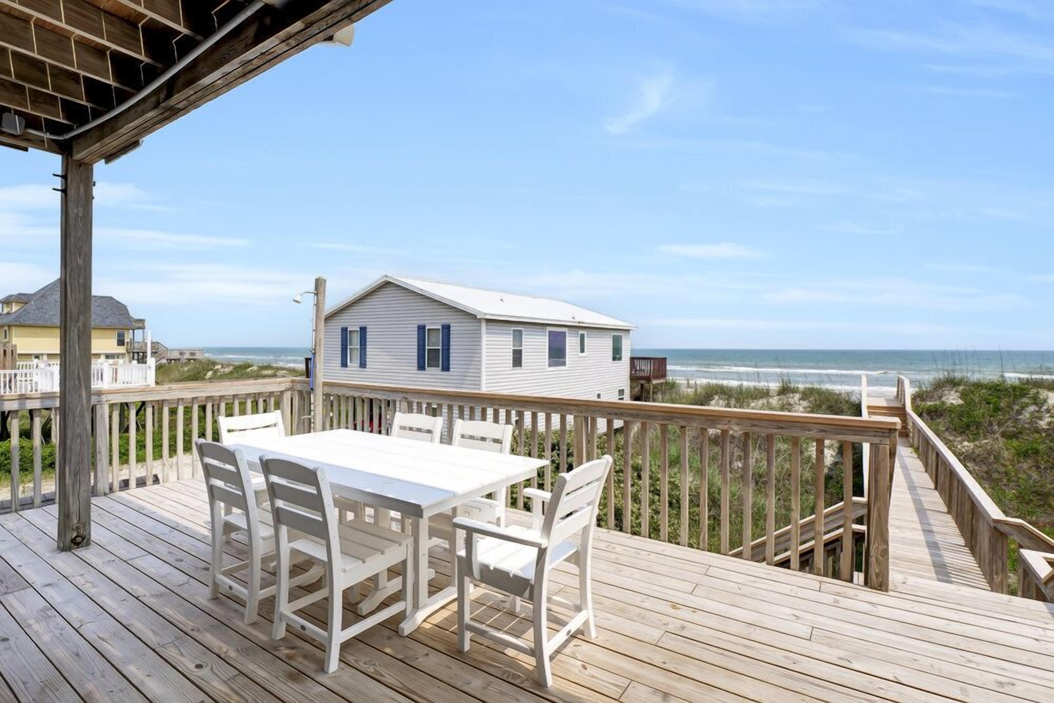 A wooden deck with a white table and chairs, overlooking a beach and a house with blue shutters.