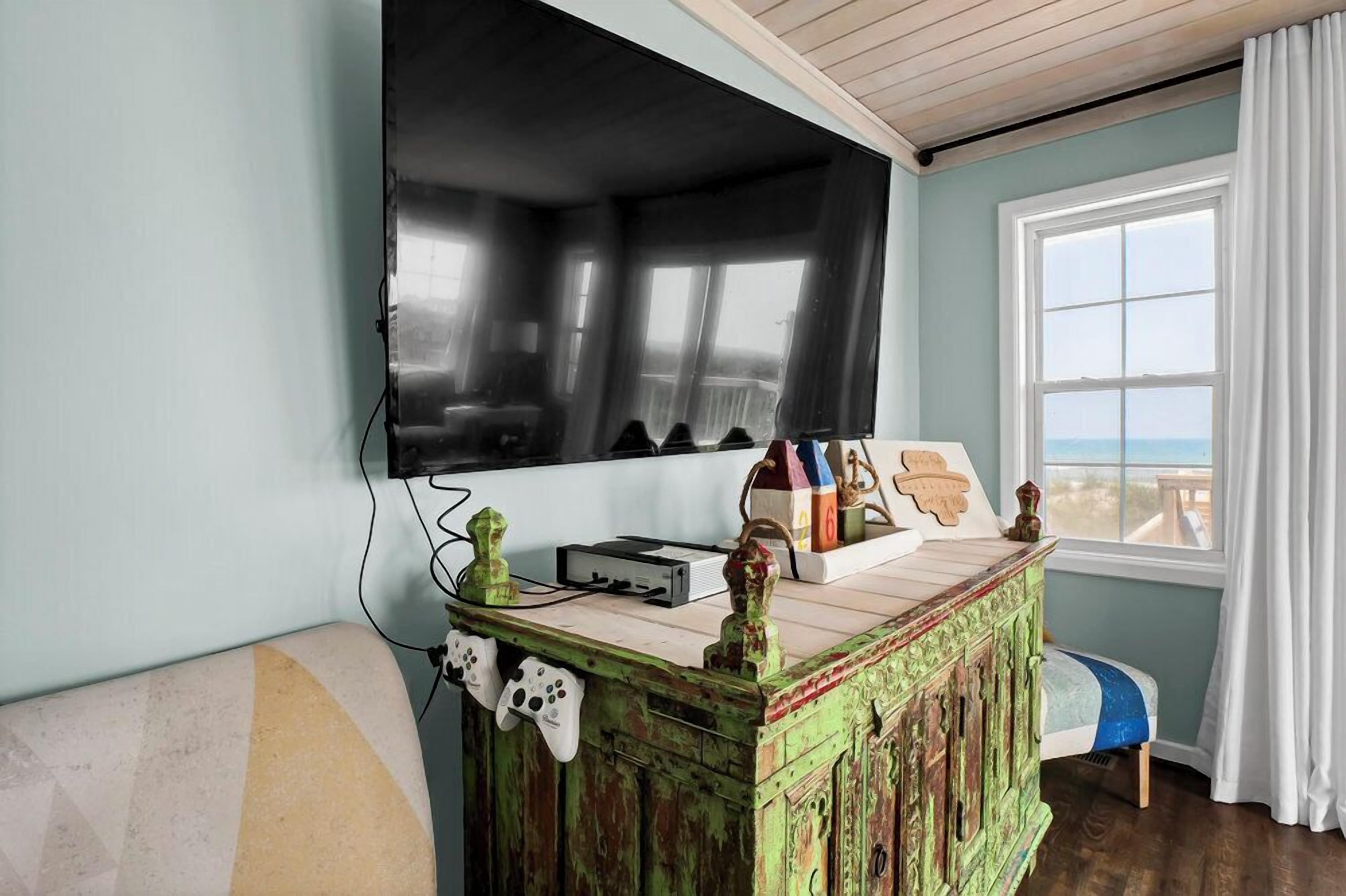 A room with a large TV mounted on the wall, a wooden cabinet with gaming controllers, and a view of the ocean through a window.