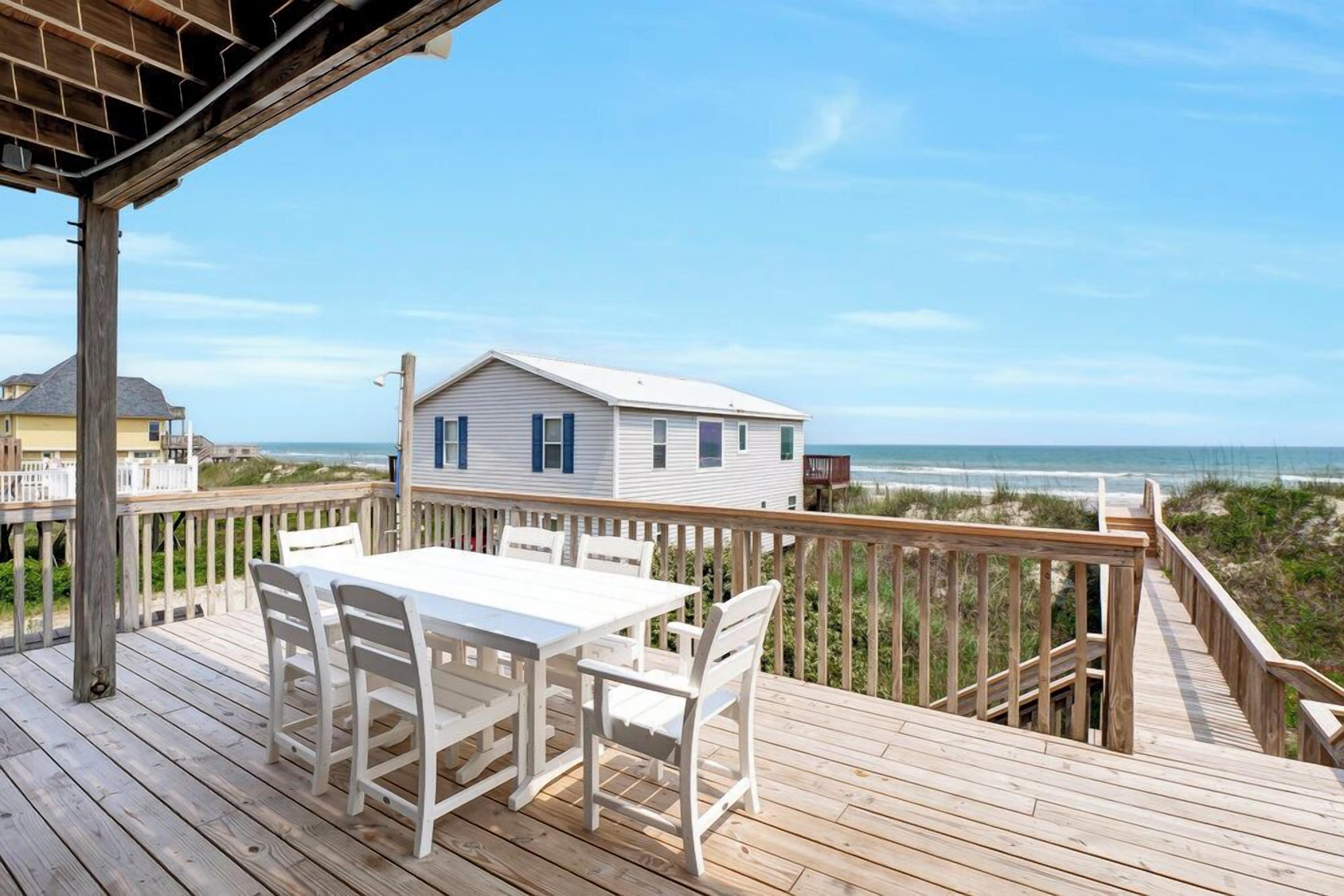 Una terraza de madera con una mesa y sillas blancas, con vistas a una playa y una casa con contraventanas azules.
