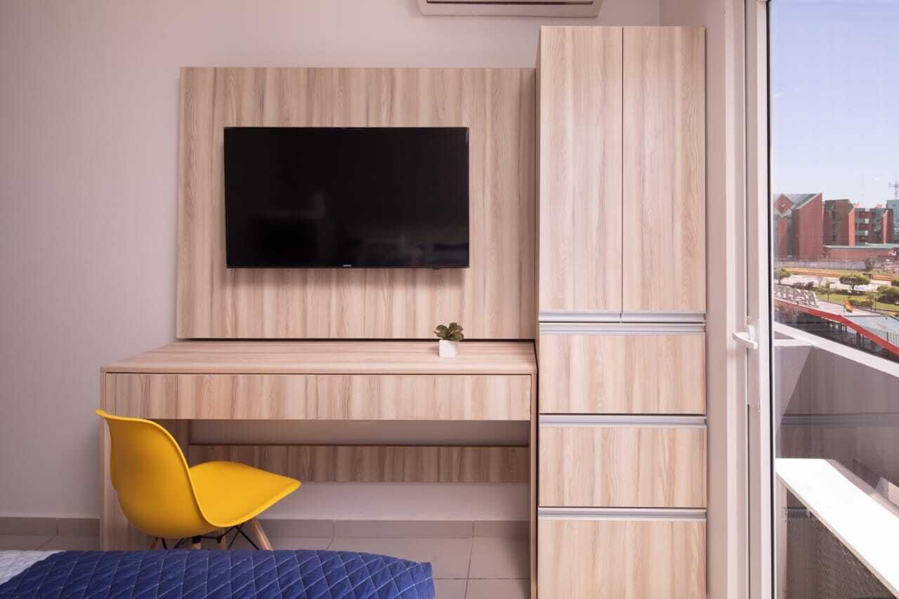 Une chambre moderne avec un téléviseur fixé au mur, un bureau, une chaise jaune et un lit recouvert d’une couverture bleue.