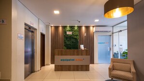 A hotel reception area with a reception desk, a waiting area with a chair and ottoman, and an elevator. - Equipetrol Suites Apart Hotel (Santa Cruz)
