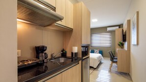 A compact kitchen with a stove, sink, and coffee maker. A bed with a blue pillow is visible in the background. - Equipetrol Suites Apart Hotel (Santa Cruz)
