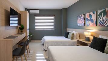 Habitación de hotel moderna con dos camas, un escritorio y sillas. Hay una ventana con persianas, un televisor de pantalla plana montado en la pared y un aire acondicionado instalado en la pared.