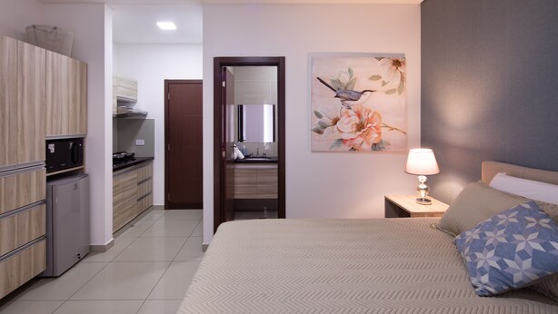 A modern hotel room with a bed, a nightstand, a lamp, a painting, a kitchen area, and a bathroom. - Equipetrol Suites Apart Hotel (Santa Cruz)