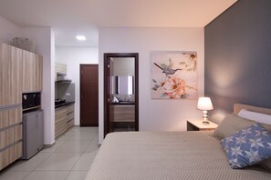 A modern hotel room with a bed, a nightstand, a lamp, a painting, a kitchen area, and a bathroom. - Equipetrol Suites Apart Hotel (Santa Cruz)