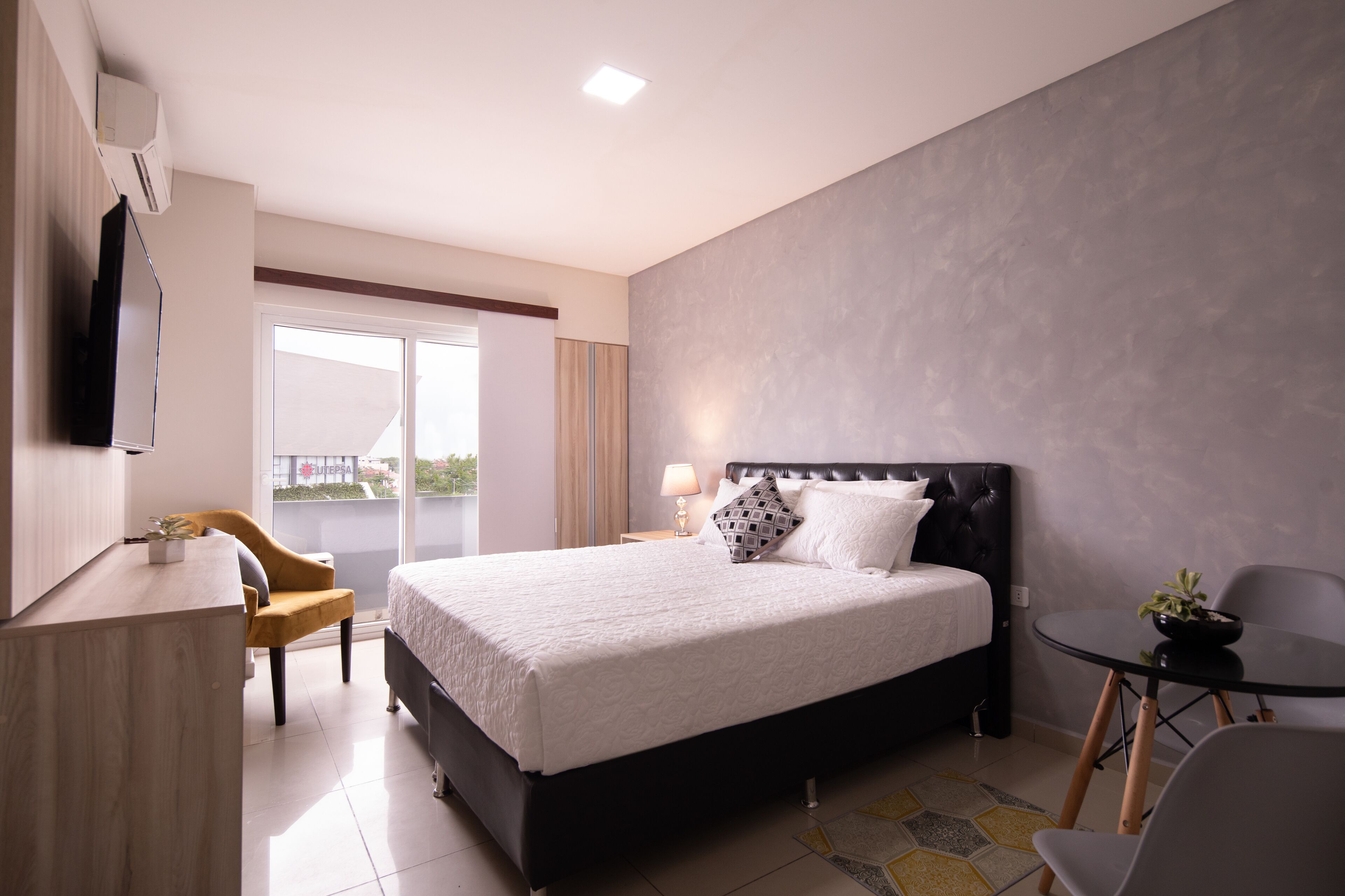 A modern hotel room with a large bed, a desk, a chair, a small table, and a balcony with a view.