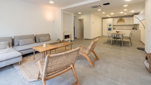 A modern living room with a sofa, a coffee table, and wicker chairs. There is a dining area with a table and chairs. The kitchen is visible in the background with modern appliances. - Newly built house with private pool (Begur)
