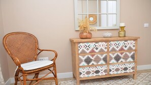 Uma cadeira de vime com almofada branca, um armário de madeira com azulejos estampados e um vaso com plantas secas em um parapeito.