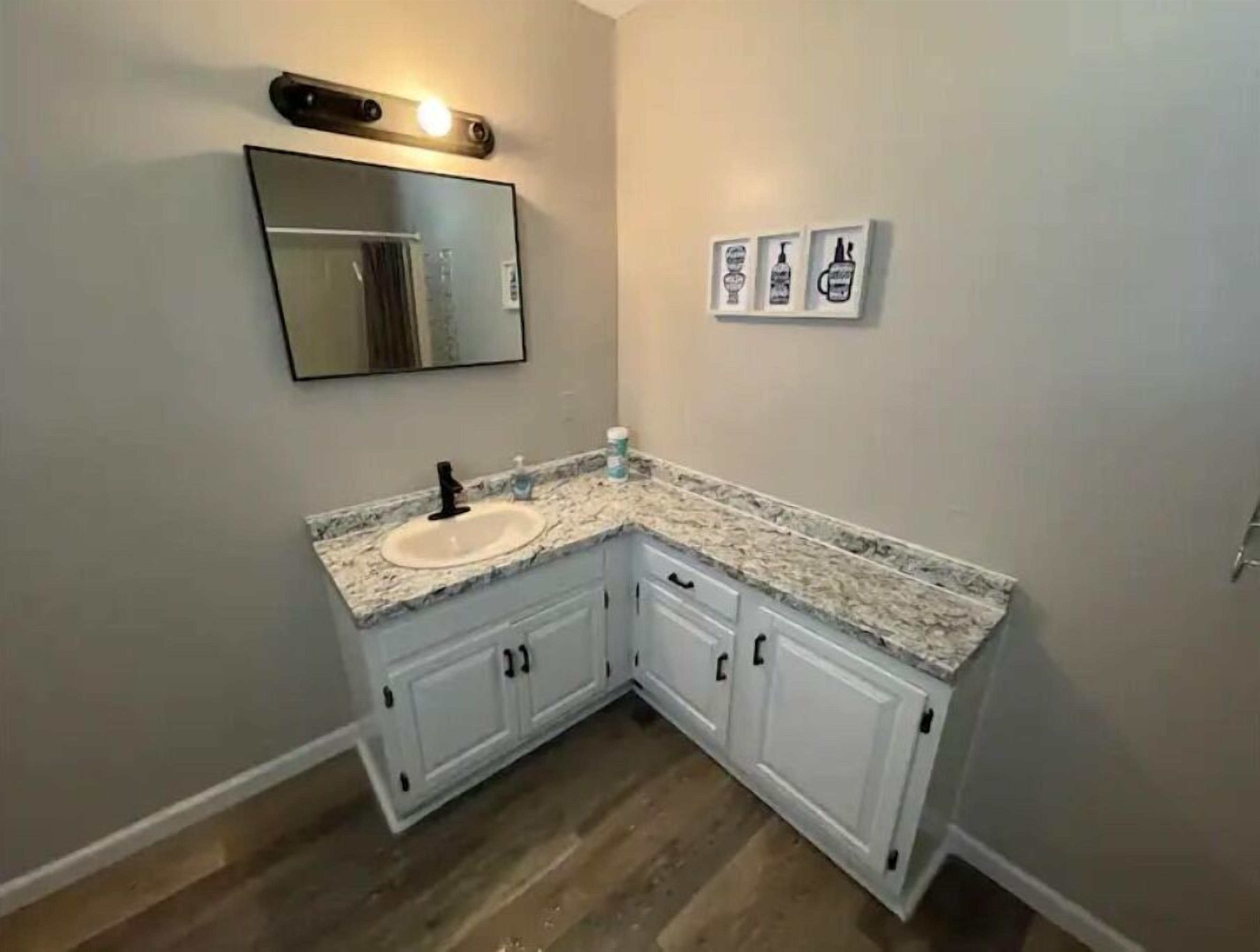 A corner bathroom with a granite countertop, white cabinets, a mirror, and three framed pictures on the wall.