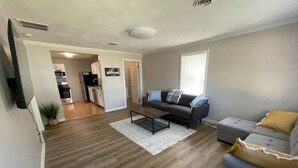 A modern living room with a grey sofa, a coffee table, and a grey loveseat. There is a kitchen in the background with a 'Welcome' sign.
