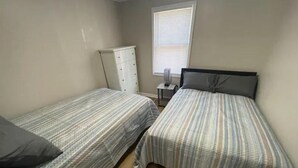 A room with two single beds, a dresser, a window with blinds, and a small table with a lamp.