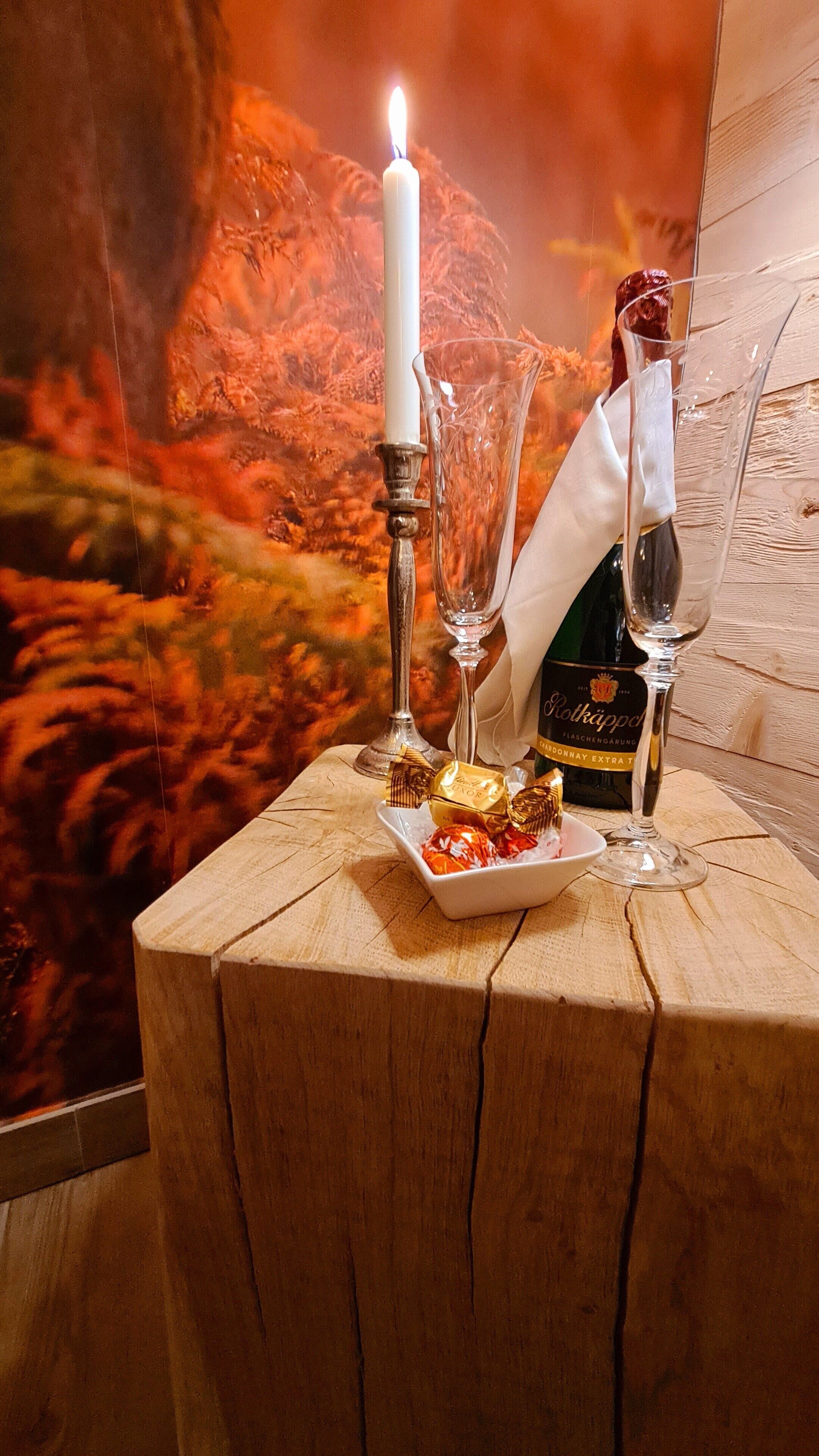 A wooden table with a bottle of wine, two wine glasses, a candle, and a plate of chocolates.