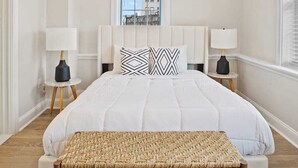 Un dormitorio con una cama grande, dos mesitas de noche con lámparas, una ventana con persianas y vistas a edificios.