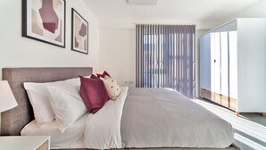 A modern bedroom with a large bed, a nightstand, and a window with vertical blinds. - Contemporary 2 Bedroom Apartment in Ashford (Ashford)