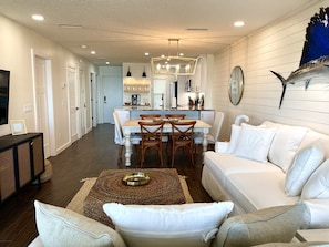 A modern living room with a white sofa, a dining area with a wooden table, and a kitchen with white cabinets. - DIRECT Oceanfront, 2BR/2BA w/Panoramic Views! 7 mins to downtown St. Augustine! (St. Augustine)