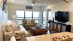 Una sala de estar con sofá, mesa de centro, mesa de comedor y televisor. Hay un balcón con vistas al océano.