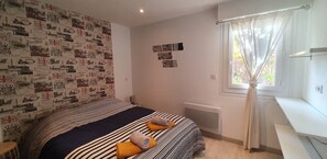 A bedroom with a bed, a radiator, a window with curtains, and a wall with vintage London-themed wallpaper. - Appartement LE Silene (Sainte-Maxime)
