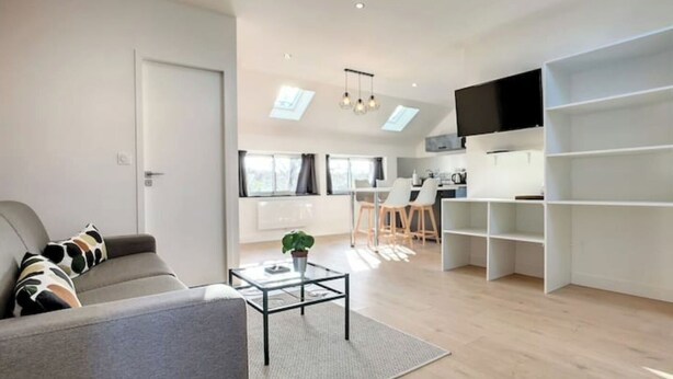 A modern living room with a grey sofa, a glass coffee table, and a dining area with a kitchen in the background.