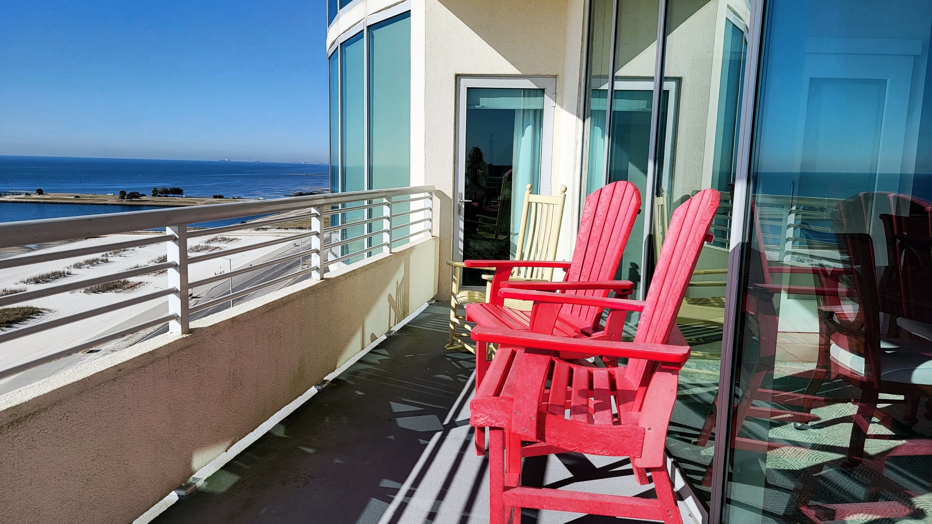 Un balcon avec des fauteuils Adirondack rouges, une porte vitrée donnant sur une pièce et une vue sur l’océan.