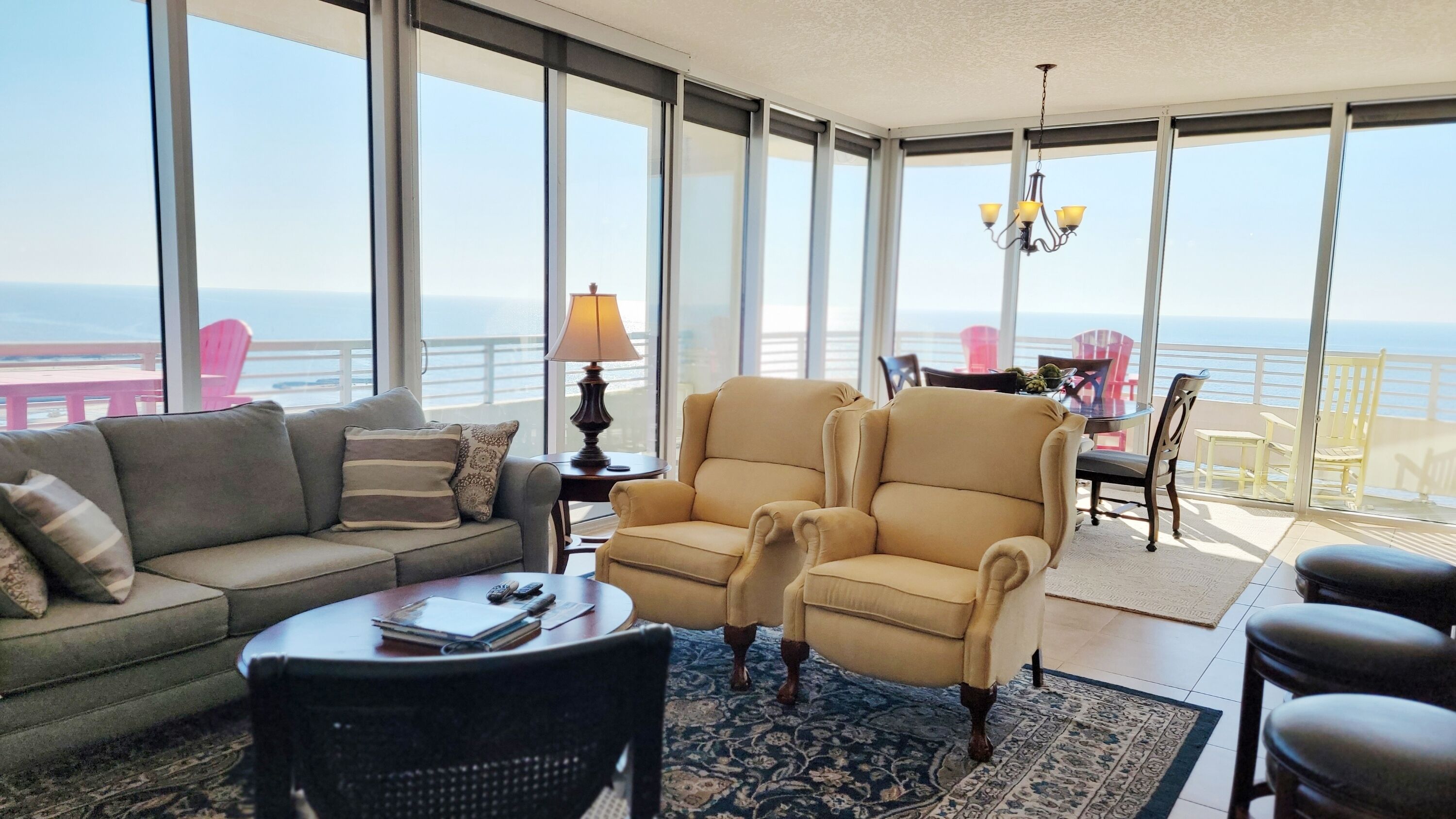 A spacious living room with a large window overlooking the ocean, a sofa, two armchairs, a small table, and a chandelier.
