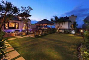 Relax in a serene outdoor space with a lawn, pathway, and seating area surrounded by lush greenery and shaded structures.