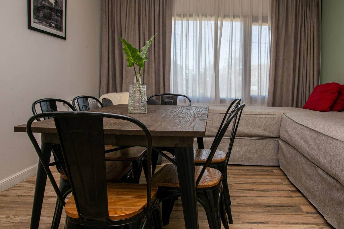 Un comedor con una mesa y sillas de madera, un florero con una planta y un sofá con cojines rojos.
