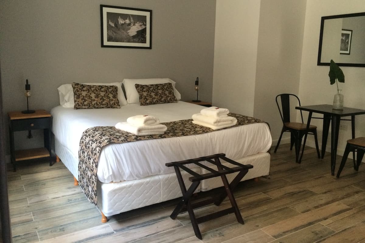 Una cama bien tendida con sábanas blancas y almohadas estampadas, una mesita de noche con una lámpara, un taburete de madera y un escritorio con una silla.