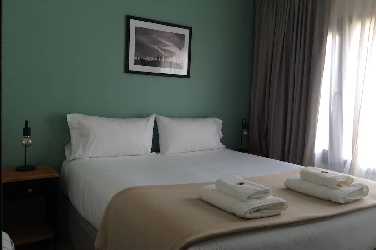 Una habitación de hotel con una cama, una mesita de noche, una lámpara, un cuadro enmarcado en la pared y una ventana con cortinas.