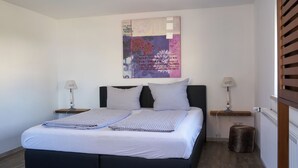 Una cama bien hecha con ropa de cama a cuadros, un cuadro abstracto grande encima y dos lámparas de noche sobre mesitas de madera.