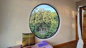 Una habitación con un ventanal grande y circular que ofrece vistas a un bosque. Hay una cama con una colcha estampada y dos almohadas. Un equipo de aire acondicionado está montado en la pared sobre el ventanal.