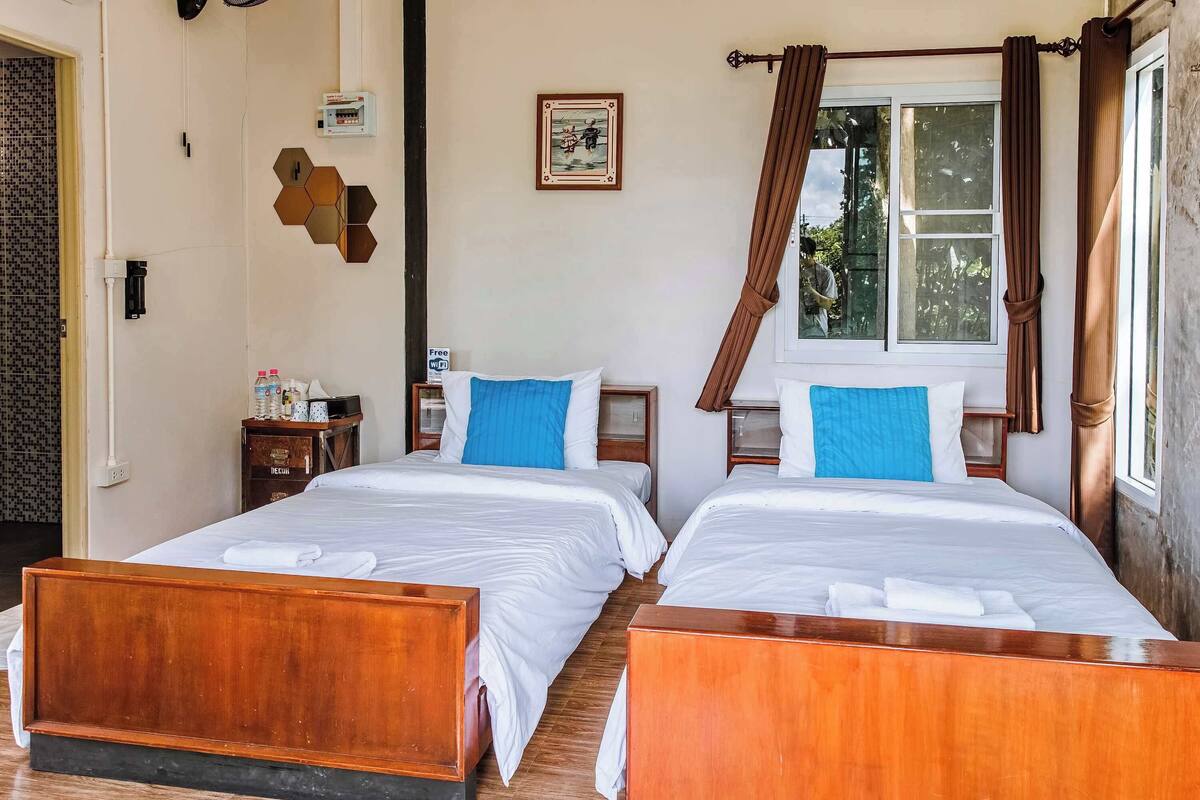 Habitación de hotel con dos camas de madera, una ventana con cortinas, un cuadro en la pared y una mesita con una botella de agua y una lámpara.