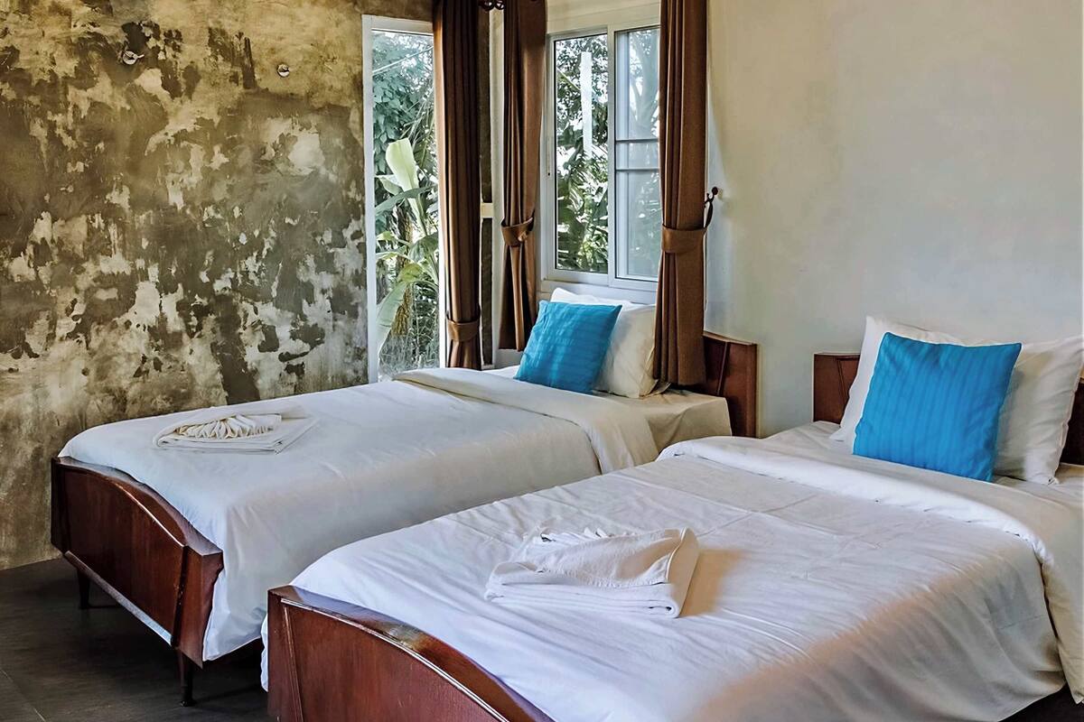 Dos camas individuales con sábanas blancas y almohadas azules, cabeceros de madera y una ventana con cortinas.