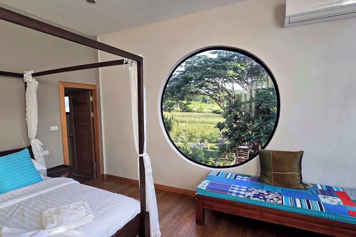 Un dormitorio con una cama con dosel, un ventanal redondo con vistas a un jardín, y una cama para el día cubierta con un edredón colorido.
