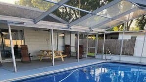 Piscina com superfície de água azul cristalina, rodeada por um deck de concreto e uma área de varanda coberta com cadeiras e uma mesa.