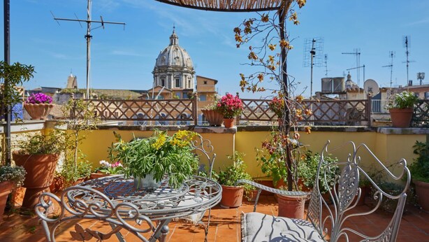 Une terrasse sur le toit, avec une table et des chaises en métal, entourée de plantes en pot et offrant une vue sur un bâtiment historique.